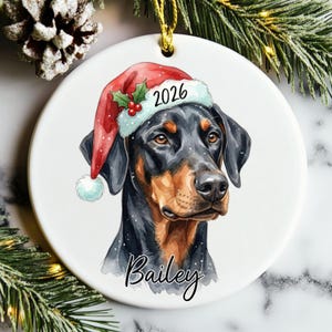 May include: A round white ceramic ornament featuring a watercolor illustration of a black and tan Doberman dog wearing a red Santa hat with holly and the year 2026. The name Bailey is printed in black script below the dog, with a gold hanging cord.