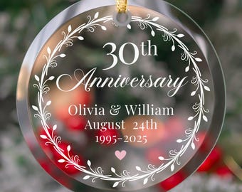 May include: Clear, round ornament with gold ribbon. The ornament features the text "30th Anniversary" with the names "Olivia & William", the date "August 24th", and the years "1995-2025". A white floral design encircles the text, with a pink heart at the bottom.