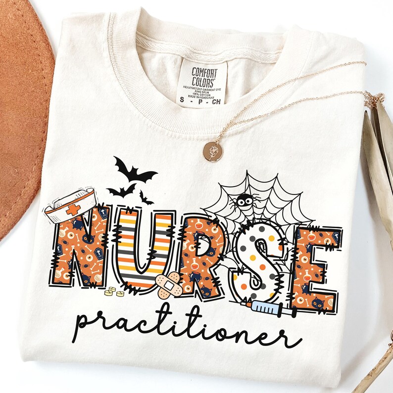 May include: Cream-colored t-shirt with a Halloween-themed design. The shirt features the words "NURSE Practitioner" in orange and black, with bats, a spider web, and medical-themed graphics. A gold necklace with a pendant is draped on the shirt.