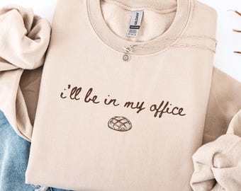 May include: A beige sweatshirt with the phrase "I'll be in my office" printed in brown script, along with a bread graphic. A silver necklace with a sunflower pendant is on the sweatshirt. The sweatshirt is folded on a white surface.
