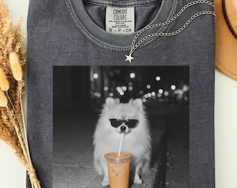 May include: A charcoal gray crewneck t-shirt with a Comfort Colors label at the inner neckline. The center features a black and white graphic of a fluffy dog wearing sunglasses and drinking from a plastic cup with a straw against a blurred background.