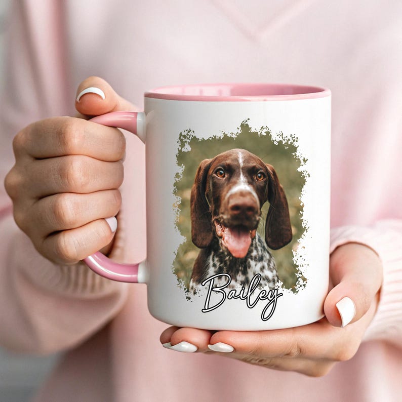 May include: White ceramic mug with a pink interior and handle. The mug features a photo of a brown and white dog with the name "Bailey" in cursive. The dog has its tongue out. The mug is held by a person.