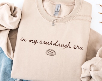 May include: A beige sweatshirt with the phrase "in my sourdough era" printed in brown script, along with a drawing of a loaf of bread. A silver necklace with a sunflower pendant rests on the shirt. The sweatshirt is folded with a pair of blue jeans.