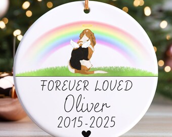 May include: A white ceramic ornament with a gold string for hanging. It features a dog illustration with angel wings and a halo, set against a rainbow. Text reads "FOREVER LOVED Oliver 2015-2025" with a heart symbol.