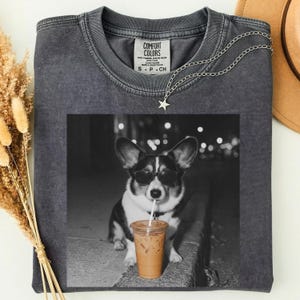 May include: A charcoal gray crew neck t-shirt featuring a black and white graphic of a Corgi wearing sunglasses and drinking from a plastic cup with a straw. The shirt has a Comfort Colors brand label at the inner neckline and a silver star necklace.