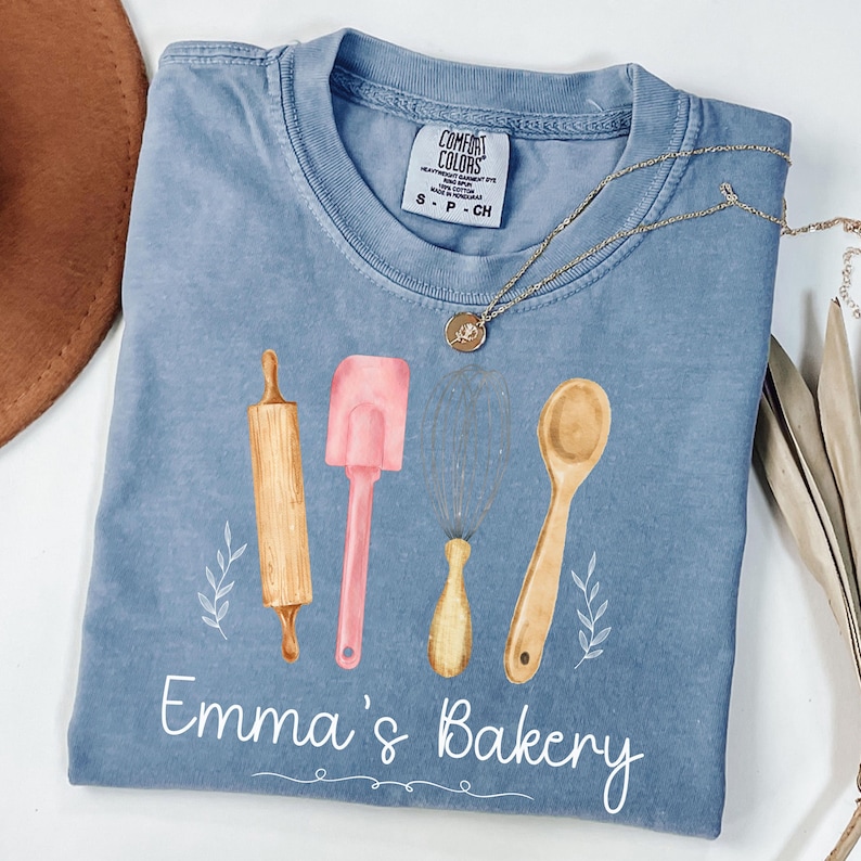 Peut inclure: Un t-shirt bleu Comfort Colors avec un motif aquarelle d'ustensiles de cuisine : un rouleau &agrave; p&acirc;tisserie, une spatule, un fouet et une cuill&egrave;re. Le texte "Emma's Bakery" est imprim&eacute; en dessous. Un collier en or est pos&eacute; sur le t-shirt.