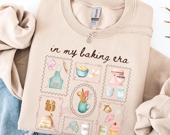 May include: A beige sweatshirt with the phrase "in my baking era" and illustrations of baking tools and ingredients. The design includes a rolling pin, apron, mixer, and cupcake. A silver necklace is draped across the top.