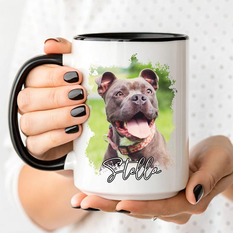 May include: White ceramic mug with a black rim and handle, featuring a portrait of a dog with its tongue out. The dog is wearing a patterned collar, and the name "Stella" is written in cursive below the image.
