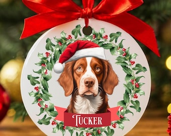 May include: A round ceramic Christmas ornament featuring a dog wearing a Santa hat, surrounded by a holly wreath. The dog's name, "Tucker," is displayed on a red banner. A red ribbon is attached for hanging.