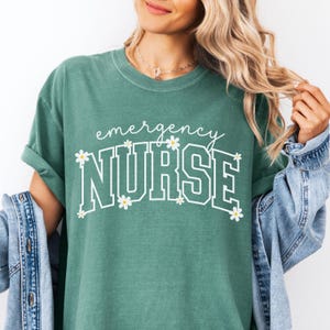 May include: A teal green t-shirt with the words "emergency NURSE" in white, decorated with small daisy flowers. The shirt has a relaxed fit and rolled-up sleeves. A denim jacket is draped over the arm.