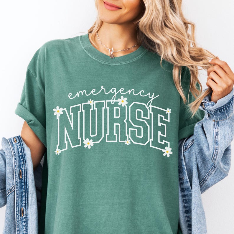 May include: A teal green t-shirt with the words "emergency NURSE" in white, decorated with small daisy flowers. The shirt has a relaxed fit and rolled-up sleeves. A denim jacket is draped over the arm.
