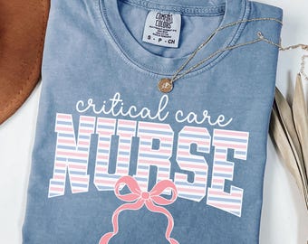 May include: A blue Comfort Colors t-shirt with the words "critical care NURSE" in white, pink, and blue stripes. A pink bow graphic is below the word "NURSE". A gold necklace with a pendant is on the shirt.