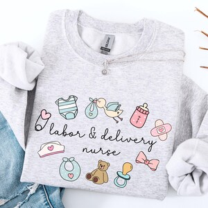 May include: A light gray sweatshirt with the text "Labor & Delivery Nurse" in black lettering. The sweatshirt features a variety of baby-related illustrations, including a baby onesie, a baby bottle, a pacifier, a teddy bear, a bow, and a stork carrying a baby.