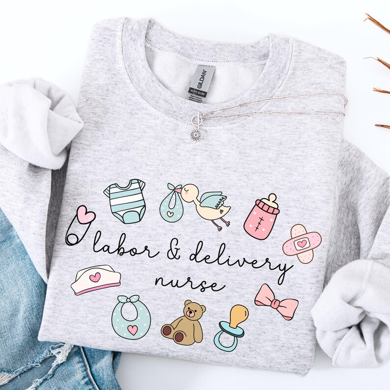 May include: A light gray sweatshirt with the text "Labor & Delivery Nurse" in black lettering. The sweatshirt features a variety of baby-related illustrations, including a baby onesie, a baby bottle, a pacifier, a teddy bear, a bow, and a stork carrying a baby.