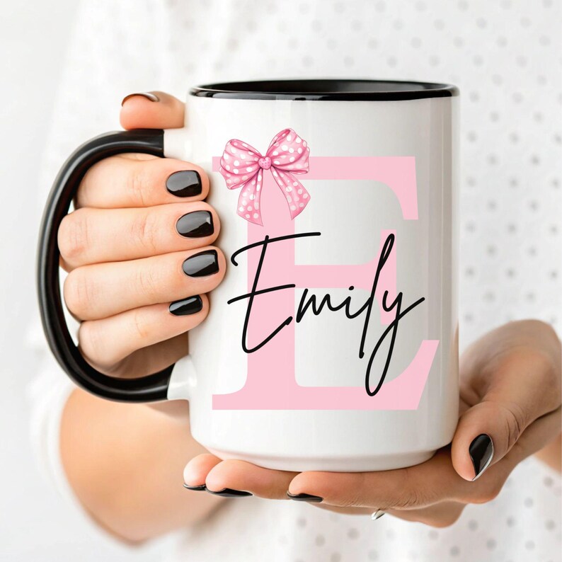 Pu&ograve; includere: Una tazza in ceramica bianca con un bordo nero. La tazza presenta una lettera "E" rosa con un fiocco rosa a pois in cima. Il nome "Emily" &egrave; scritto in corsivo nero sotto la lettera.