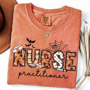 May include: Orange Comfort Colors t-shirt with a Halloween-themed "NURSE practitioner" design. The design includes bats, a spider web, and medical-themed illustrations. A gold necklace with a pendant is draped across the shirt.