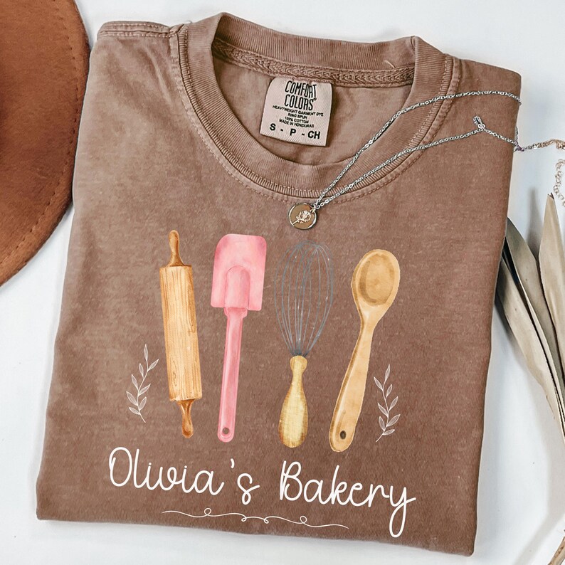 Peut inclure: Un t-shirt marron Comfort Colors avec un motif aquarelle d'ustensiles de cuisine : un rouleau &agrave; p&acirc;tisserie, une spatule, un fouet et une cuill&egrave;re. Le texte "Olivia's Bakery" est imprim&eacute; en dessous. Un collier en argent avec un pendentif est pos&eacute; sur le t-shirt.