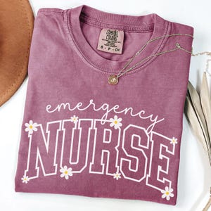 May include: A mauve-colored t-shirt with the words "emergency NURSE" in white, decorated with small daisy graphics. A gold necklace with a pendant rests on the shirt. The shirt is folded and lies on a white surface.