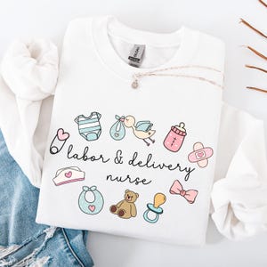 May include: White sweatshirt with the text "labor & delivery nurse" and various baby-related icons, including a baby bottle, a pacifier, a baby onesie, a bib, a teddy bear, a bow, and a safety pin.
