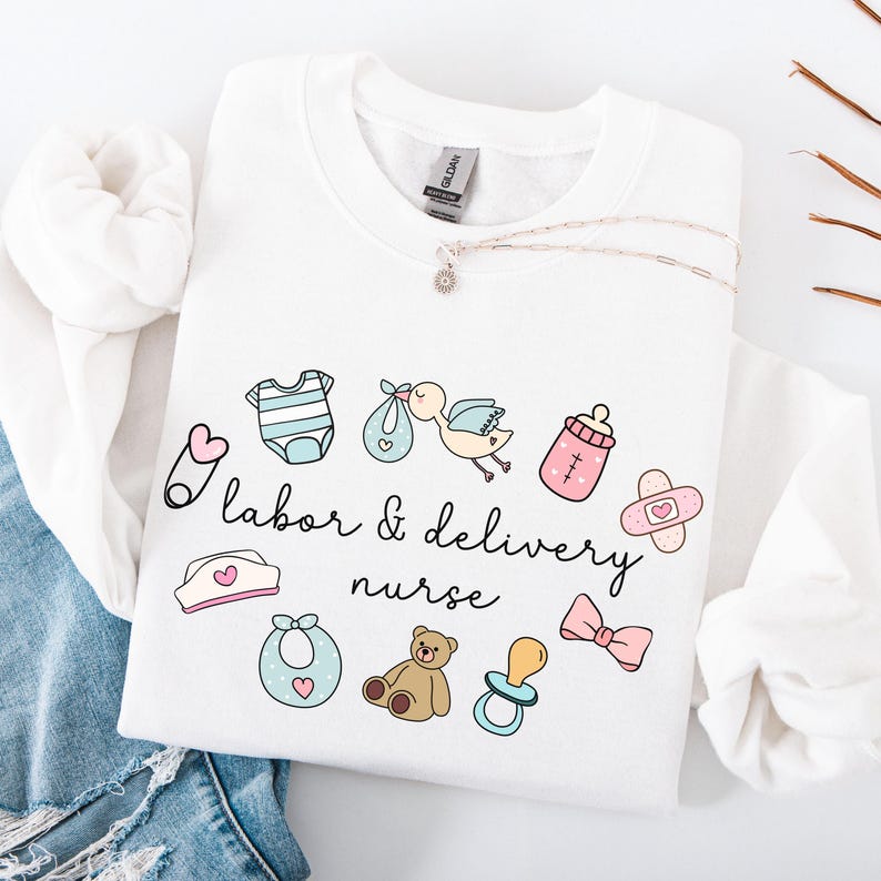 May include: White sweatshirt with the text "labor & delivery nurse" and various baby-related icons, including a baby bottle, a pacifier, a baby onesie, a bib, a teddy bear, a bow, and a safety pin.