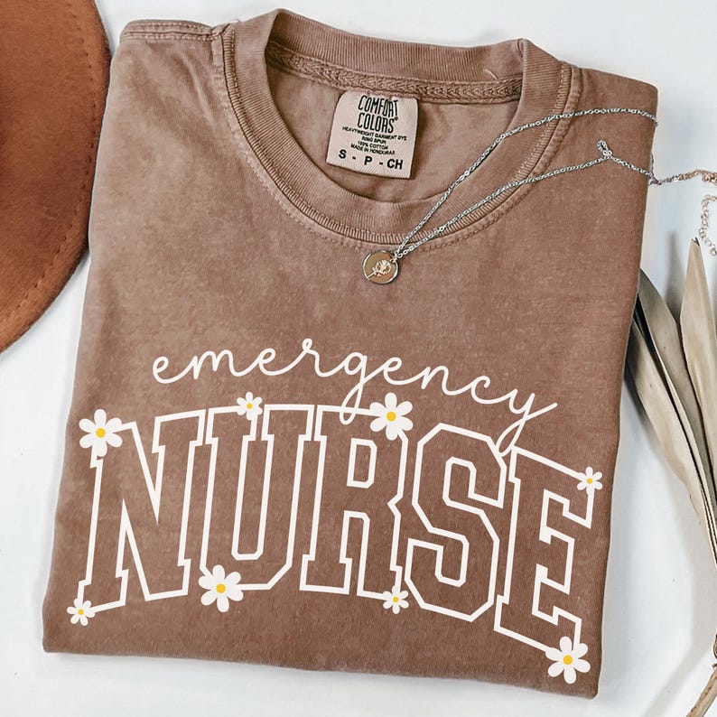 May include: A brown Comfort Colors t-shirt with the words "emergency NURSE" in white, decorated with daisy graphics. A silver necklace with a pendant rests on the shirt. The shirt is folded and displayed on a white surface.