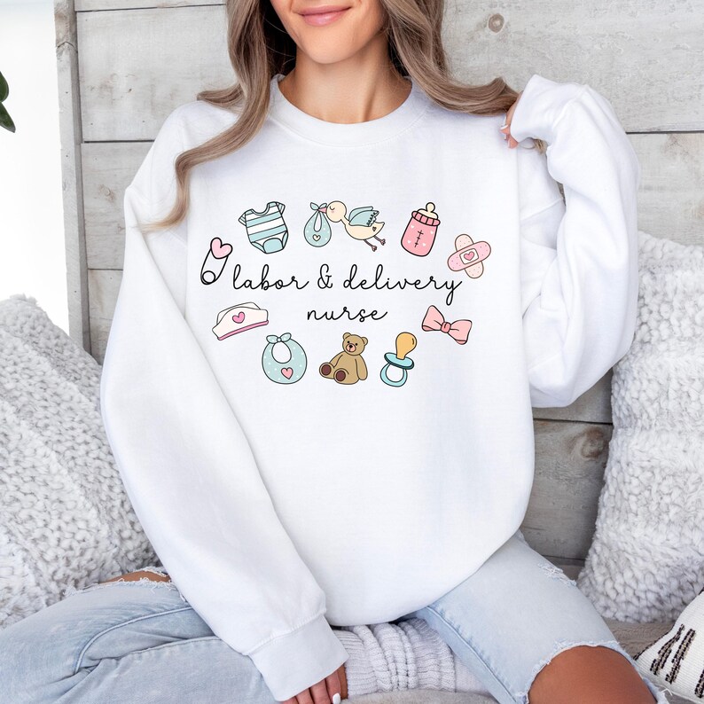 May include: White sweatshirt with a pink and blue design of baby items and the text "Labor & Delivery Nurse".