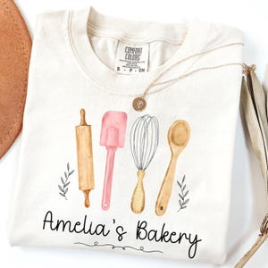 Peut inclure: T-shirt cr&egrave;me avec des illustrations &agrave; l'aquarelle d'ustensiles de cuisine : un rouleau &agrave; p&acirc;tisserie, une spatule, un fouet et une cuill&egrave;re. Le texte "Amelia's Bakery" est en dessous. Un collier en or avec un pendentif est drap&eacute; sur le t-shirt.