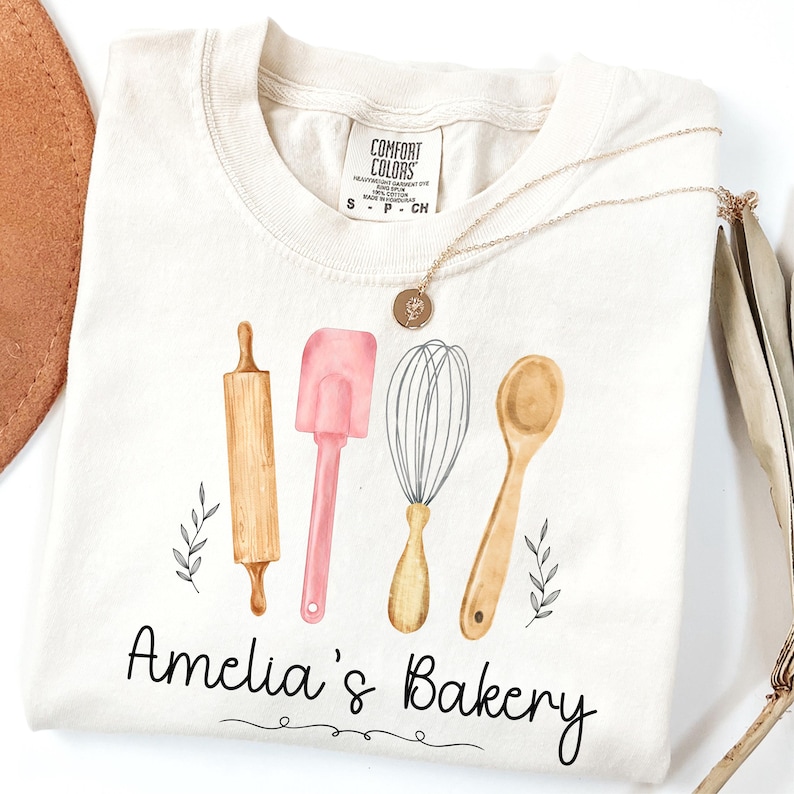 Peut inclure: T-shirt cr&egrave;me avec des illustrations &agrave; l'aquarelle d'ustensiles de cuisine : un rouleau &agrave; p&acirc;tisserie, une spatule, un fouet et une cuill&egrave;re. Le texte "Amelia's Bakery" est en dessous. Un collier en or avec un pendentif est drap&eacute; sur le t-shirt.