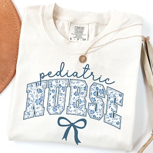 May include: A cream-colored t-shirt with a blue floral print that says "pediatric nurse" in a collegiate font with a blue bow underneath.