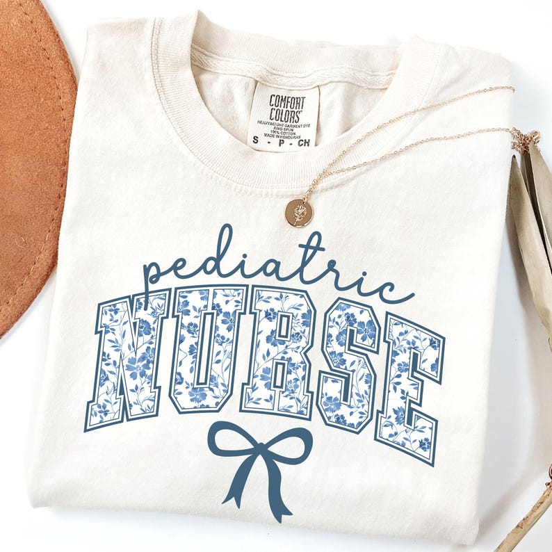 May include: A cream-colored t-shirt with a blue floral print that says "pediatric nurse" in a collegiate font with a blue bow underneath.