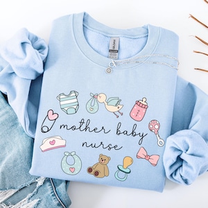 May include: Light blue sweatshirt with a graphic design of baby items, including a pacifier, a bottle, a baby onesie, a teddy bear, a bib, and a stork carrying a baby. The text "I mother baby nurse" is printed on the sweatshirt.