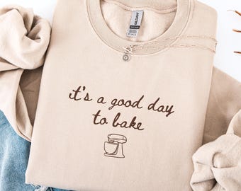 May include: Beige crewneck sweatshirt with the phrase "it's a good day to bake" in brown script, along with a mixer graphic. A silver necklace with a sunflower pendant is on the sweatshirt. The sweatshirt is folded on a white surface.