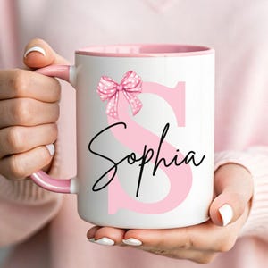 Pu&ograve; includere: Una tazza in ceramica bianca con bordo e manico rosa. La tazza presenta una lettera "S" rosa con un fiocco rosa a pois bianchi in cima. Il nome "Sophia" &egrave; scritto in corsivo nero sotto la lettera.