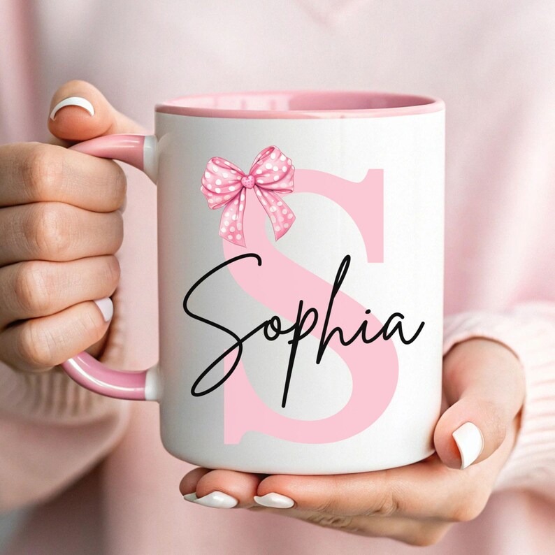 Pu&ograve; includere: Una tazza in ceramica bianca con bordo e manico rosa. La tazza presenta una lettera "S" rosa con un fiocco rosa a pois bianchi in cima. Il nome "Sophia" &egrave; scritto in corsivo nero sotto la lettera.