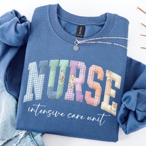 May include: A blue sweatshirt with the word "NURSE" in patchwork letters. The letters are in various pastel colors and patterns. Below the word "NURSE" is the text "intensive care unit" in a cursive font. A silver necklace with a flower pendant is on the sweatshirt.