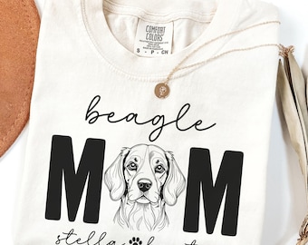 May include: Cream-colored t-shirt with the words "beagle MOM" in black block letters, a beagle illustration, and the name "stella baxter" with a paw print. A gold necklace is draped across the shirt.