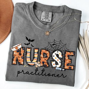 May include: A gray Comfort Colors t-shirt with a Halloween-themed design. The shirt features the word "NURSE" in orange and black, with bats, a spiderweb, and a syringe. The word "practitioner" is printed below.