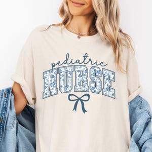May include: A cream colored t-shirt with the text "pediatric NURSE" in blue with a blue floral pattern. A blue bow is below the text.