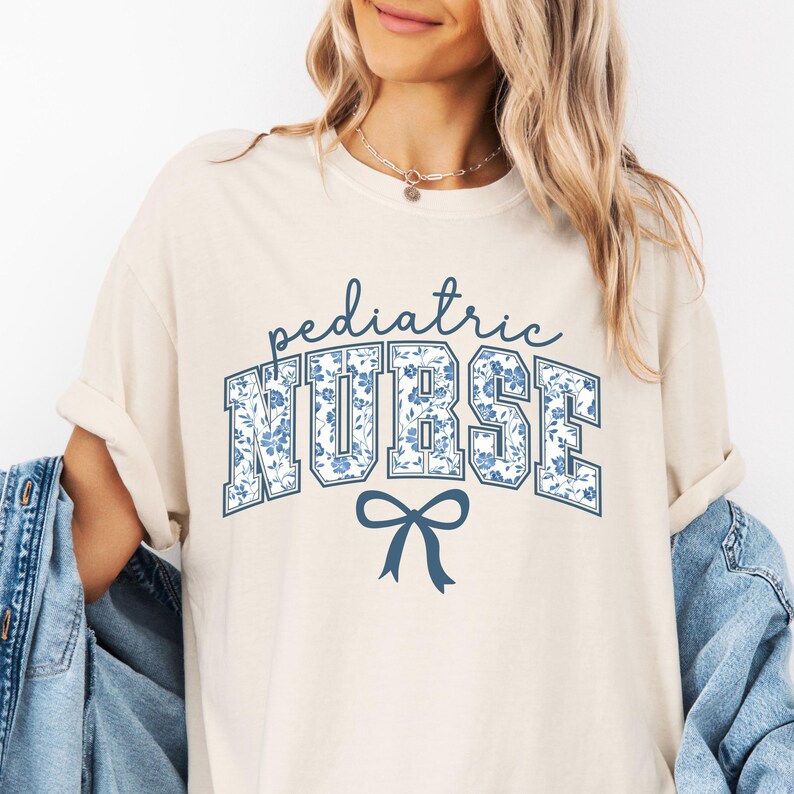 May include: A cream colored t-shirt with the text "pediatric NURSE" in blue with a blue floral pattern. A blue bow is below the text.