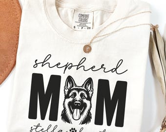 May include: Cream-colored t-shirt with the words "shepherd MOM" in black block letters, with a graphic of a German Shepherd dog. The name "stella baxter" is written below, along with a paw print. A gold necklace is draped across the shirt.