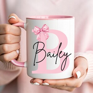 Pu&ograve; includere: Una tazza in ceramica bianca con un bordo e un manico rosa. La tazza presenta una grande lettera rosa "B" con un fiocco rosa a pois in cima. Il nome "Bailey" &egrave; scritto in corsivo nero sotto la lettera.
