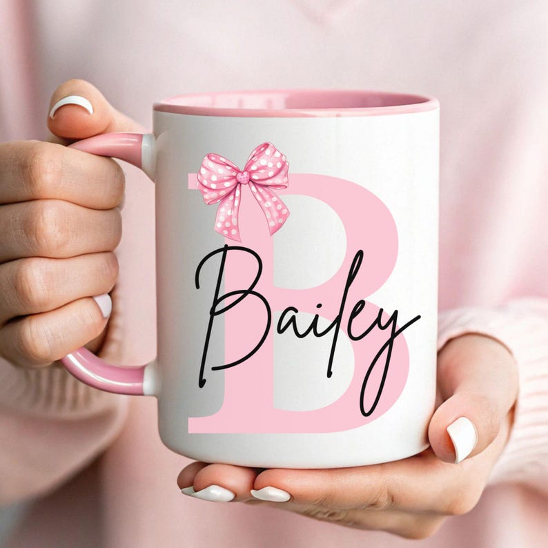 Pu&ograve; includere: Una tazza in ceramica bianca con un bordo e un manico rosa. La tazza presenta una grande lettera rosa "B" con un fiocco rosa a pois in cima. Il nome "Bailey" &egrave; scritto in corsivo nero sotto la lettera.