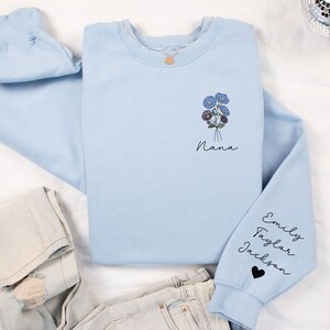 May include: Light blue sweatshirt with a floral design and the word "Nana" on the front. The sleeves have the names "Emily", "Taylor", and "Jackson" written on them.