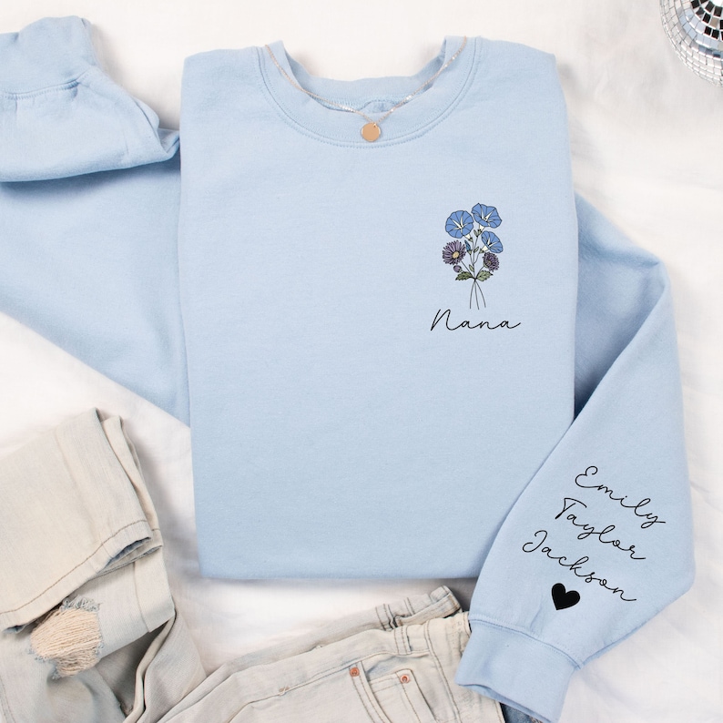 May include: Light blue sweatshirt with a floral design and the word "Nana" on the front. The sleeves have the names "Emily", "Taylor", and "Jackson" written on them.