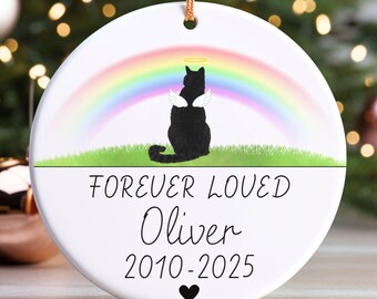 May include: A white ceramic ornament with a gold string for hanging. It features a black cat with angel wings and a halo, a rainbow, and the text "FOREVER LOVED Oliver 2010-2025" with a heart symbol. The ornament is a memorial.