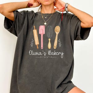 Peut inclure: T-shirt gris fonc&eacute; avec un motif aquarelle d'ustensiles de cuisine : un rouleau &agrave; p&acirc;tisserie, une spatule, un fouet et une cuill&egrave;re. Le texte "Olivia's Bakery" est &eacute;crit en dessous des outils dans une police cursive. Le t-shirt est de style d&eacute;contract&eacute;.
