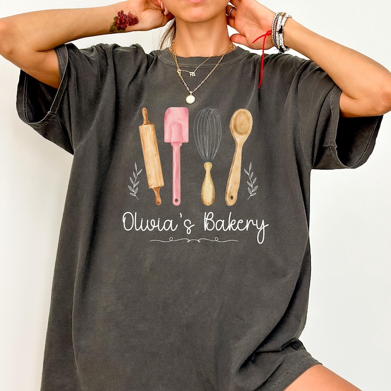 Peut inclure: T-shirt gris fonc&eacute; avec un motif aquarelle d'ustensiles de cuisine : un rouleau &agrave; p&acirc;tisserie, une spatule, un fouet et une cuill&egrave;re. Le texte "Olivia's Bakery" est &eacute;crit en dessous des outils dans une police cursive. Le t-shirt est de style d&eacute;contract&eacute;.