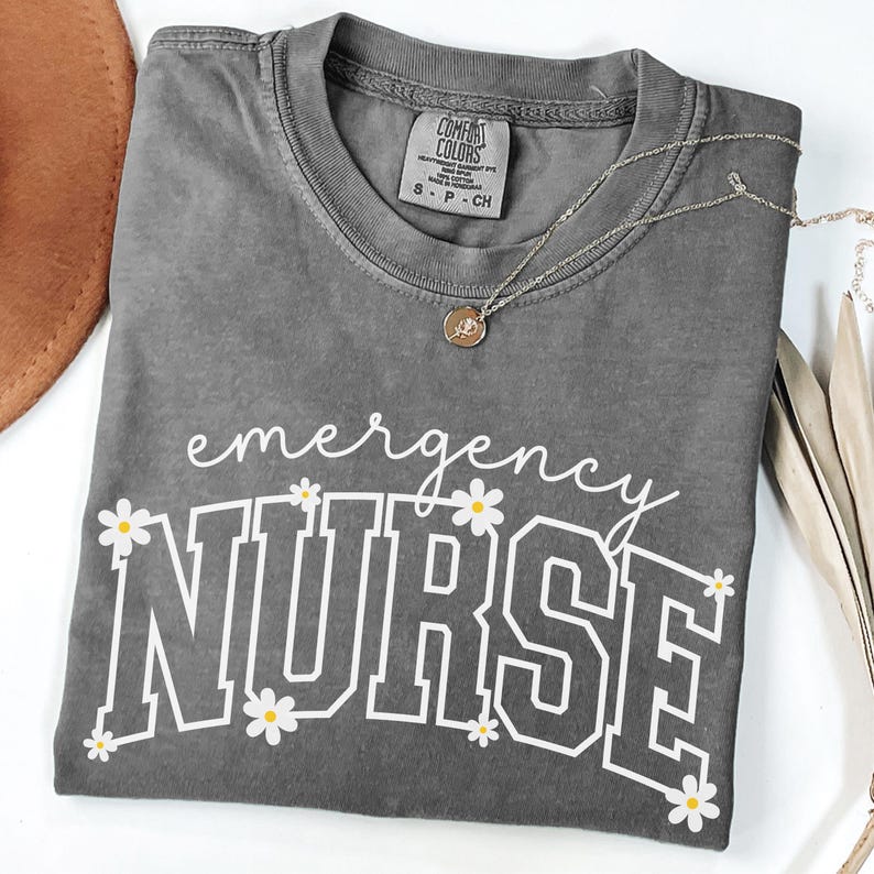 May include: A gray Comfort Colors t-shirt with the words "emergency NURSE" in white, decorated with daisy graphics. A silver necklace with a small pendant rests on the shirt. The shirt is folded on a white surface.