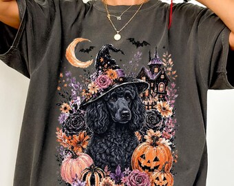 May include: A charcoal gray t-shirt featuring a graphic print of a black poodle wearing a witch hat adorned with flowers. The design includes a crescent moon, bats, a haunted house, pumpkins, and various colorful floral elements surrounding the dog.