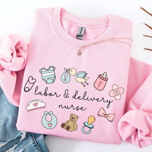 May include: Pink sweatshirt with a cute design featuring baby-related icons and the text "labor & delivery nurse".